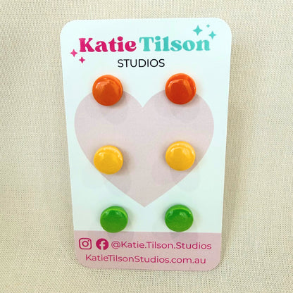 Circular stud earrings in orange, yellow and lime green displayed on a Katie Tilson Studios branded card against a beige background.