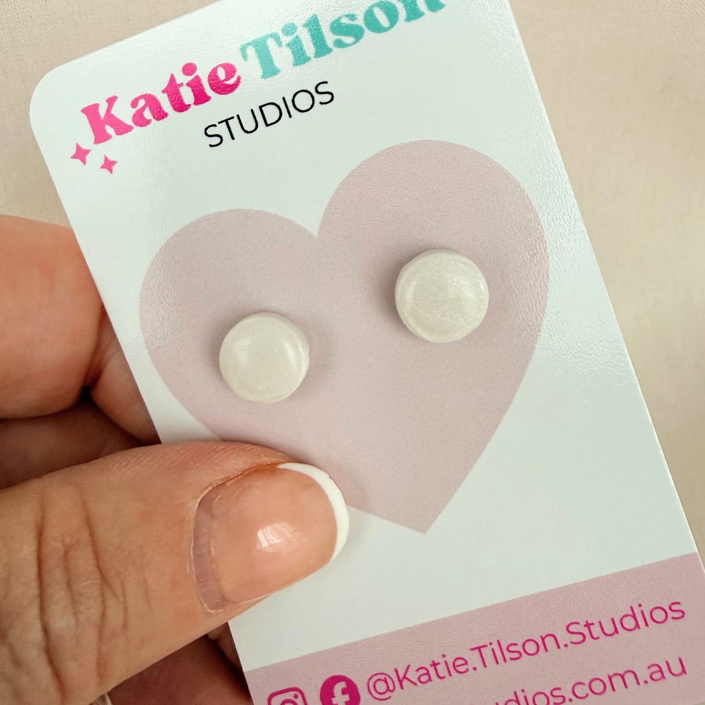 White circular stud earrings on a Katie Tilson Studios card held by a hand.