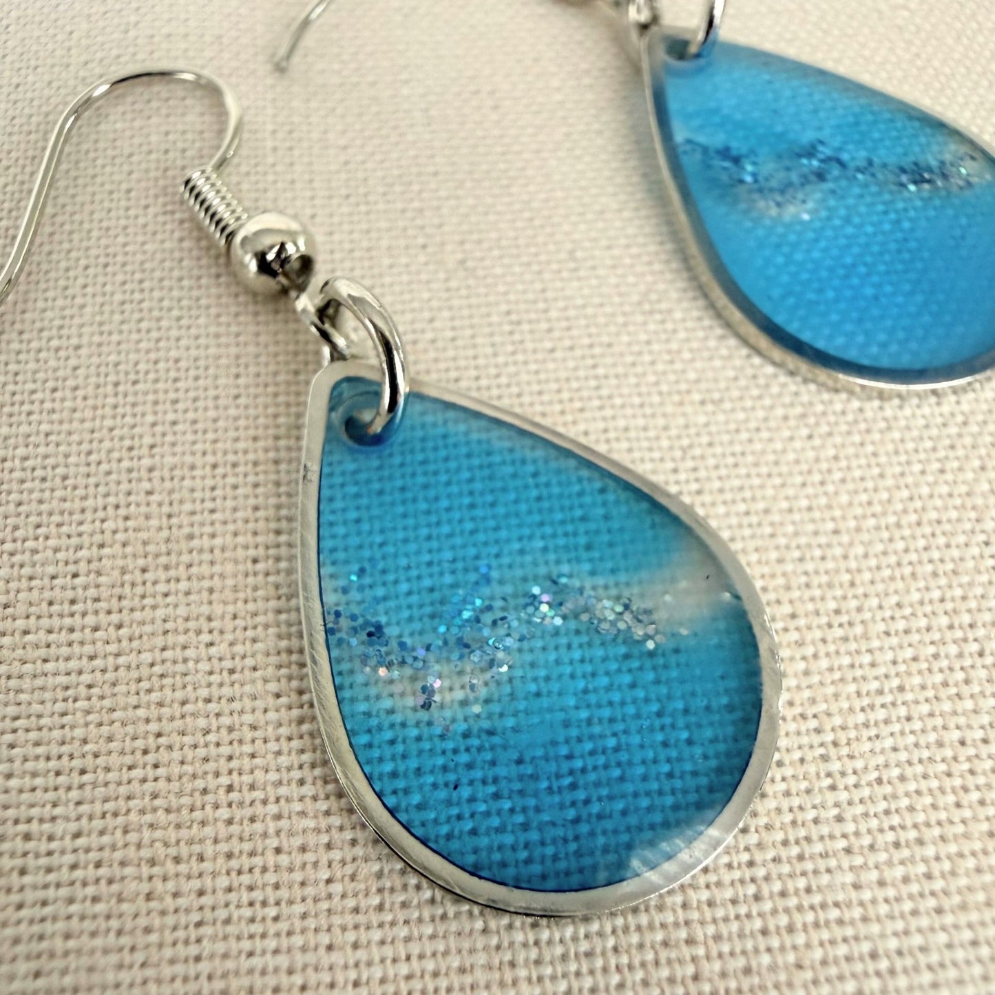 Close up of lightweight dangle earrings Coastal Glow in blue resin with silver details and findings.