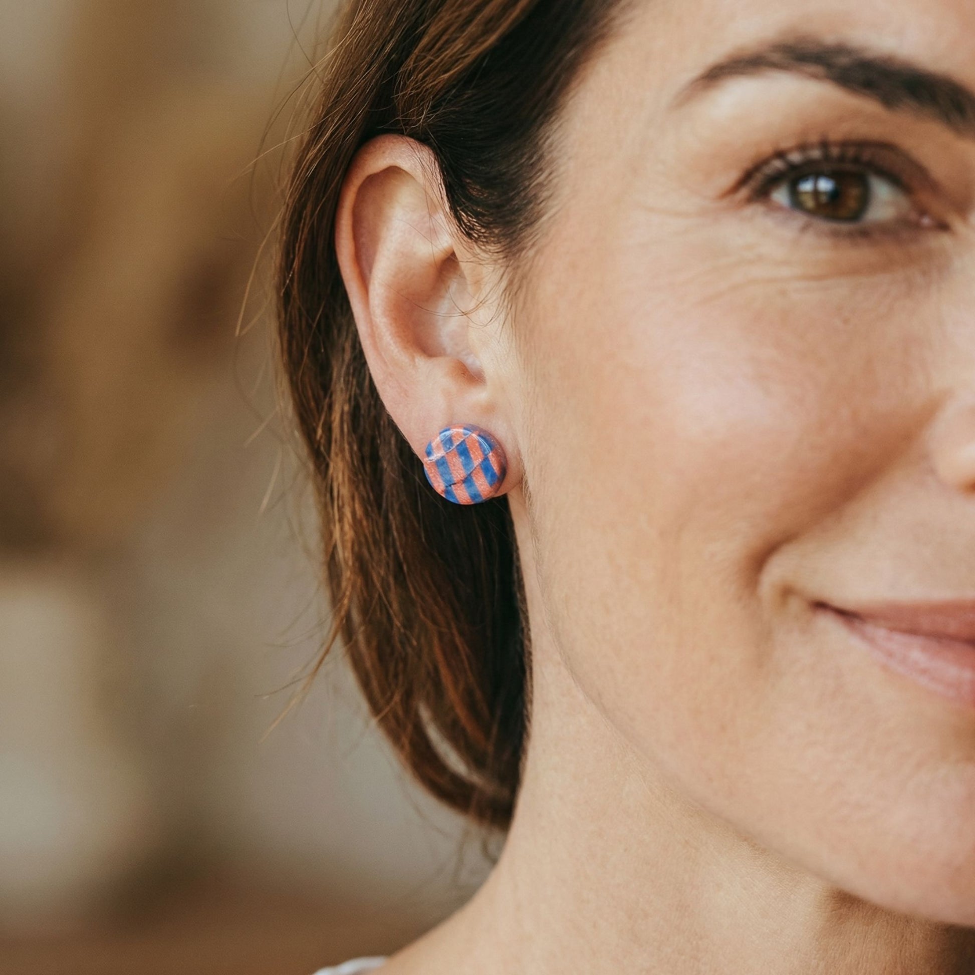 Close-up of a woman wearing colorful cobalt blue and coral checkered stud earrings with a blurred background.