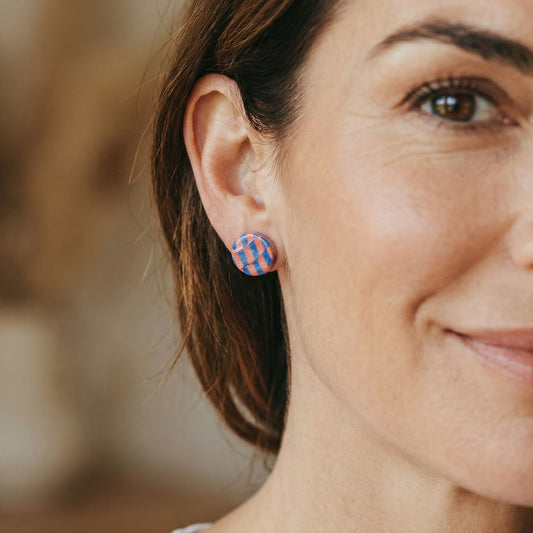 Close-up of a woman wearing colorful cobalt blue and coral checkered stud earrings with a blurred background.