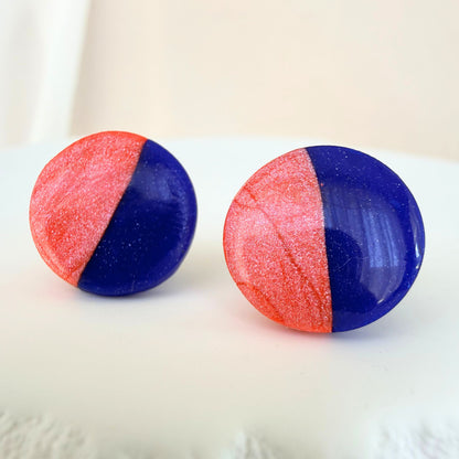 A pair of cobalt blue and coral half and half colour circle studs with shimmer and glossy resin finish resting on a white surface.