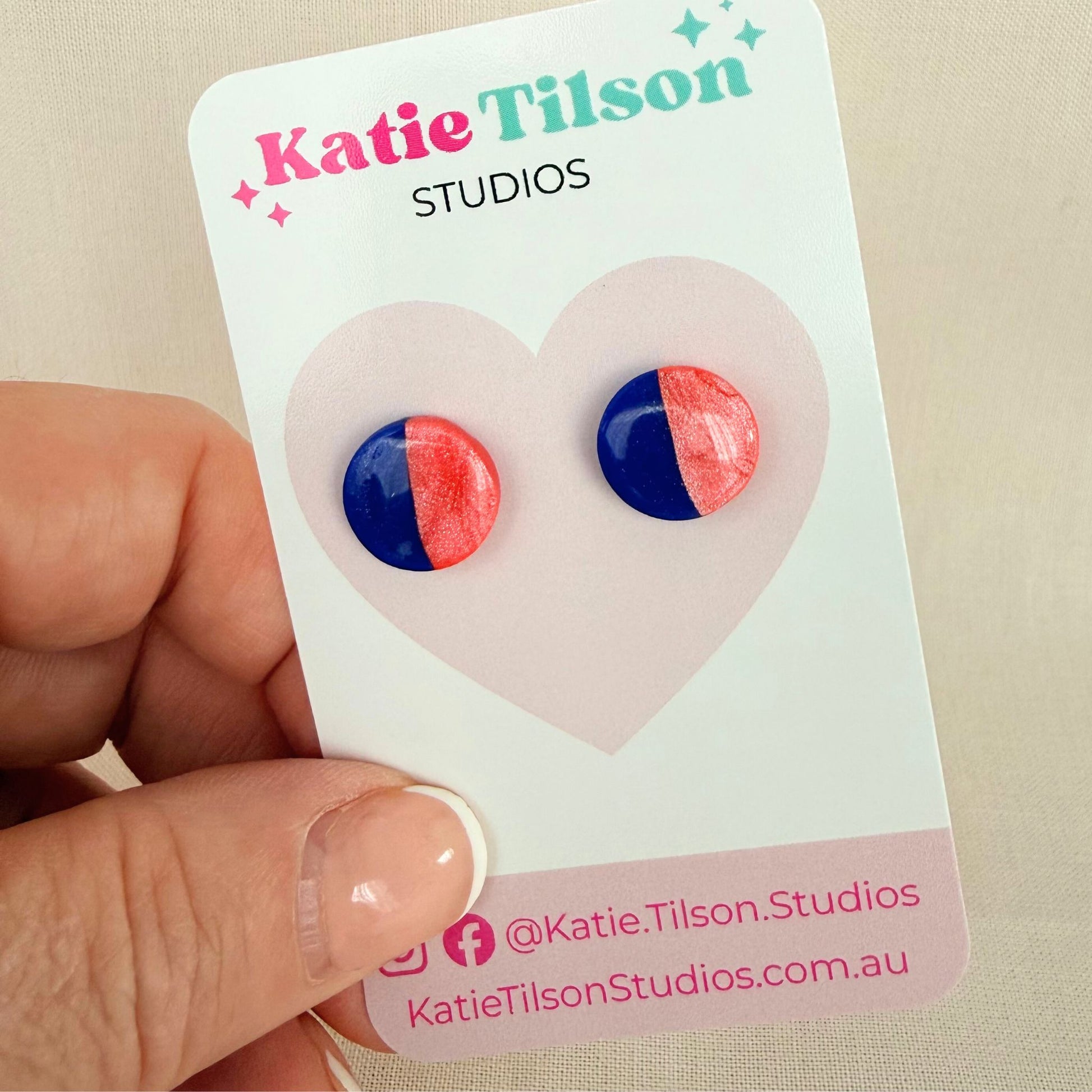Cobalt blue and coral half and half  design circular stud earrings on a branded earring card being held by a hand against a beige background.