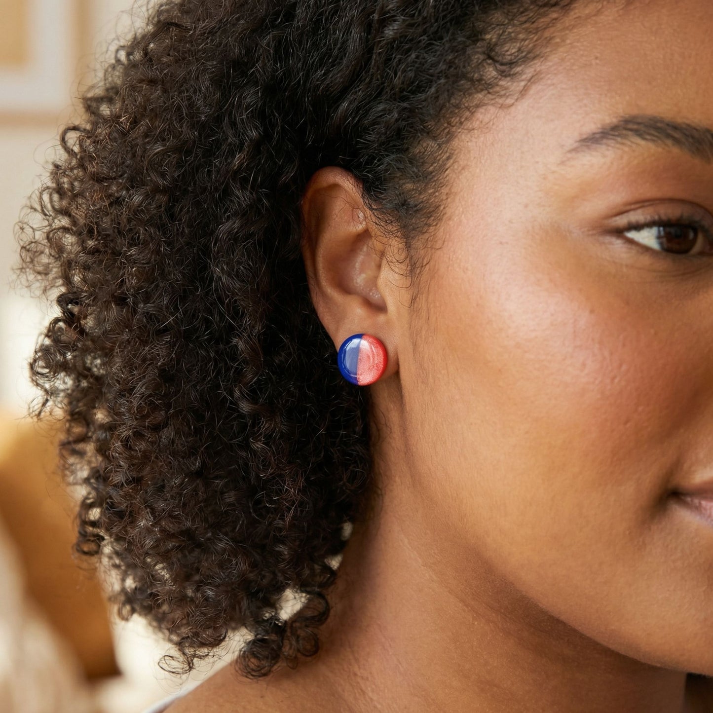 Close-up of a woman wearing round cobalt blue and coral half-and-half circular stud earrings with a glossy finish against a blurred background.