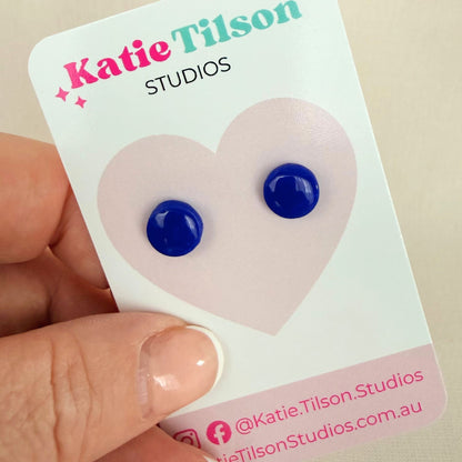 A pair of cobalt blue circular stud earrings on a branded earring card held by a hand.