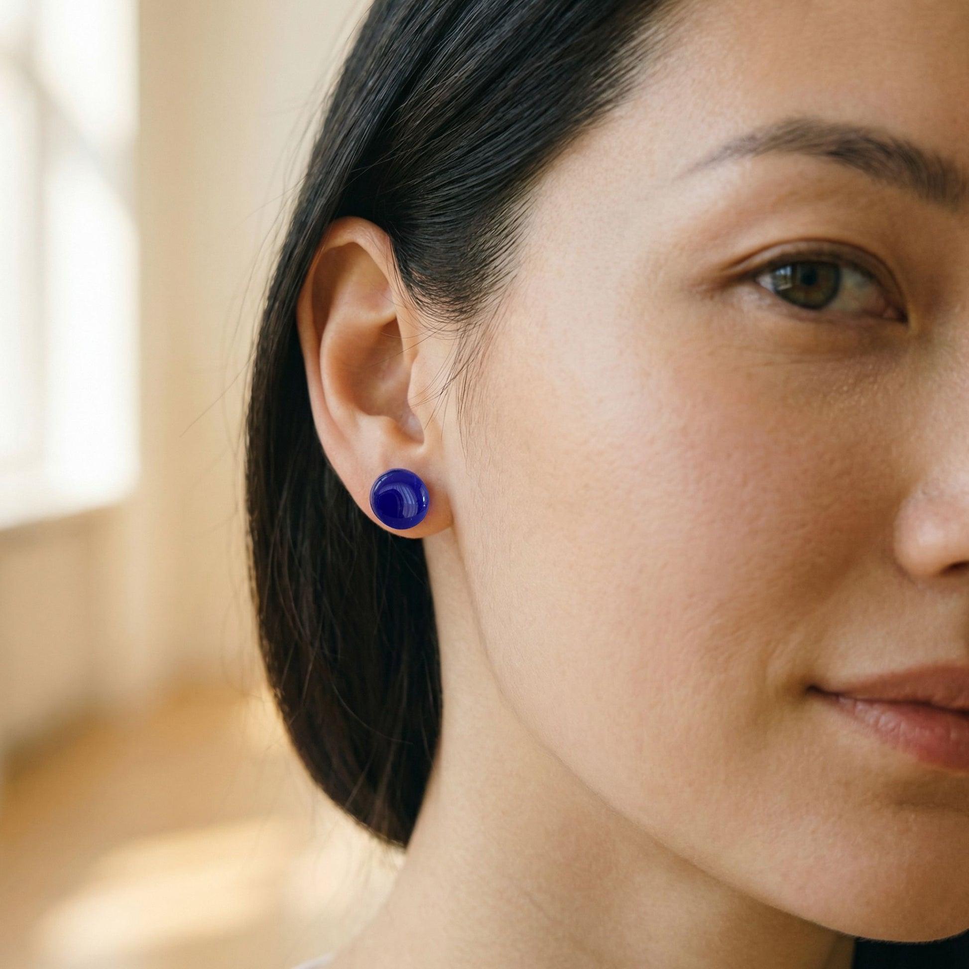 Close-up of a woman wearing round cobalt blue stud earrings with a glossy finish against a blurred background.