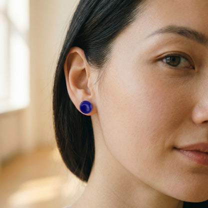 Close-up of a woman wearing round cobalt blue stud earrings with a glossy finish against a blurred background.
