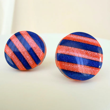 A pair of bright cobalt blue and coral stripe circle studs with shimmer and glossy resin finish resting on a white surface.