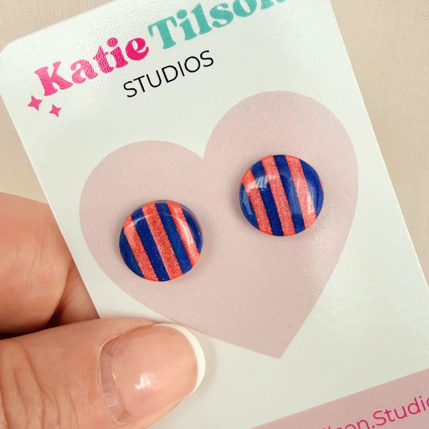 Cobalt blue and coral striped circular stud earrings on a Katie Tilson Studios card held by a hand.
