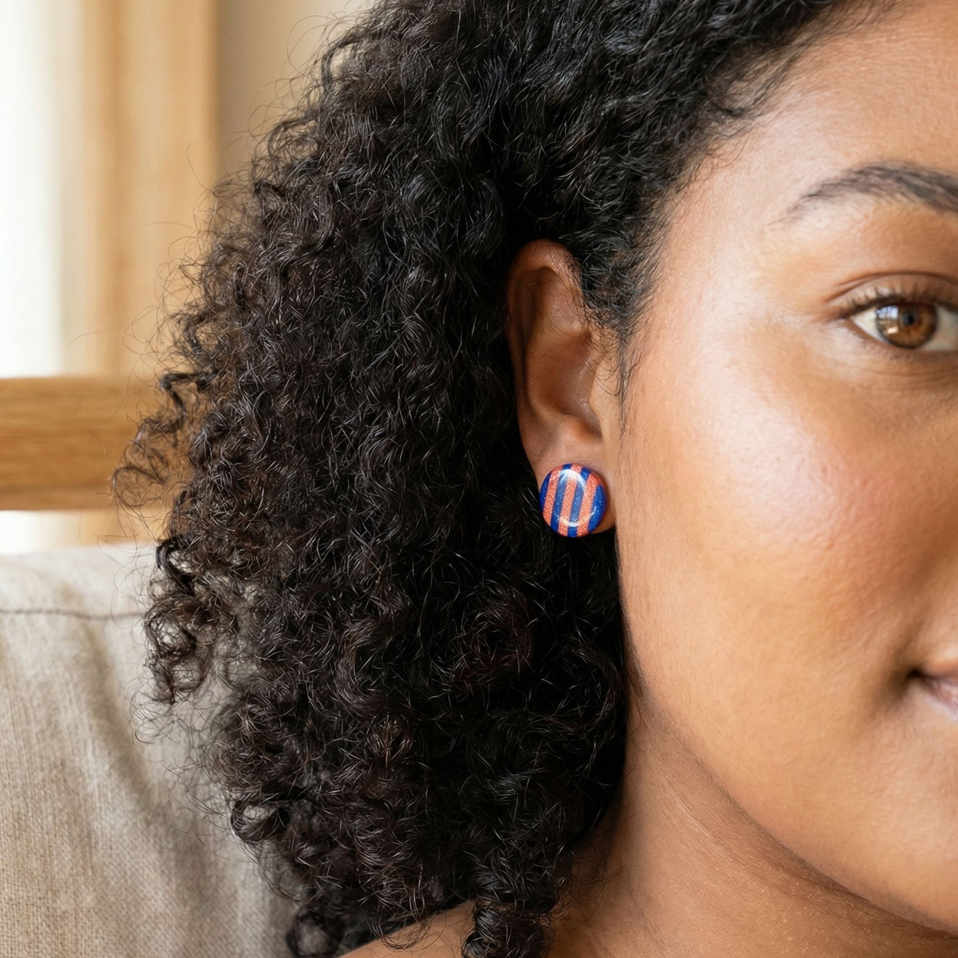 Close-up of a woman wearing colorful cobalt and coral striped stud earrings with a blurred background.