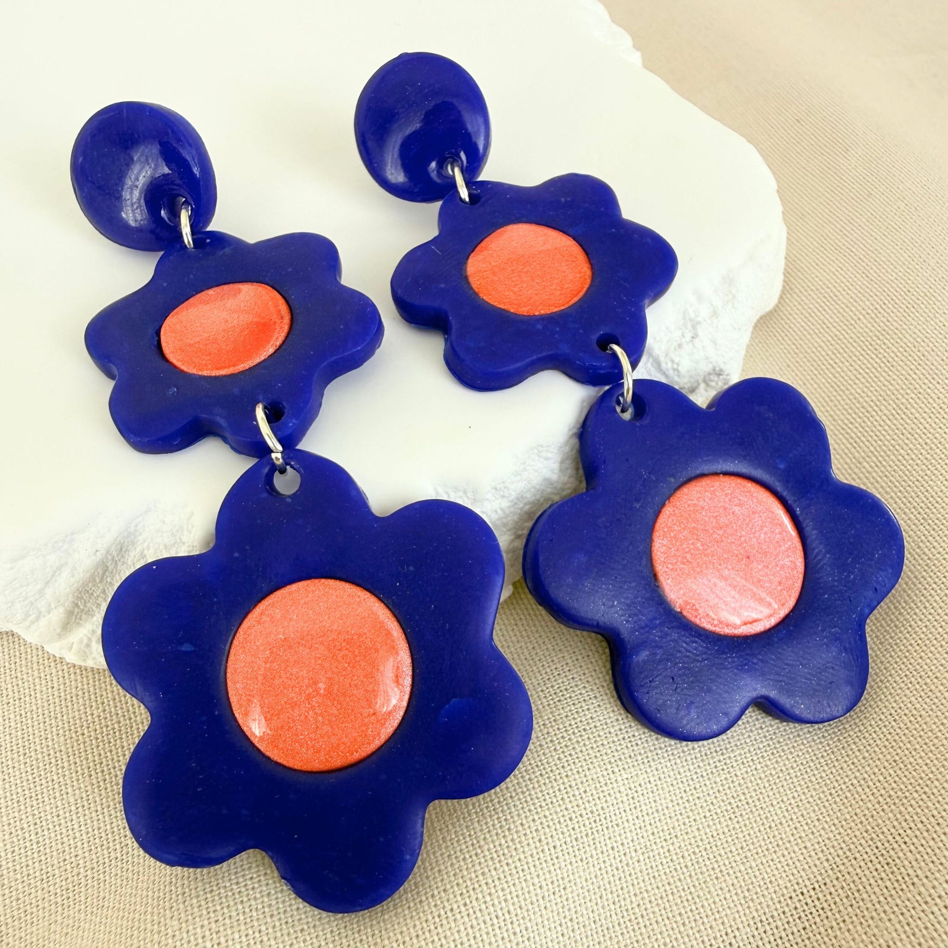 Flower-shaped 3 tier drop earrings with cobalt blue petals and glossy coral centers on a beige fabric background.
