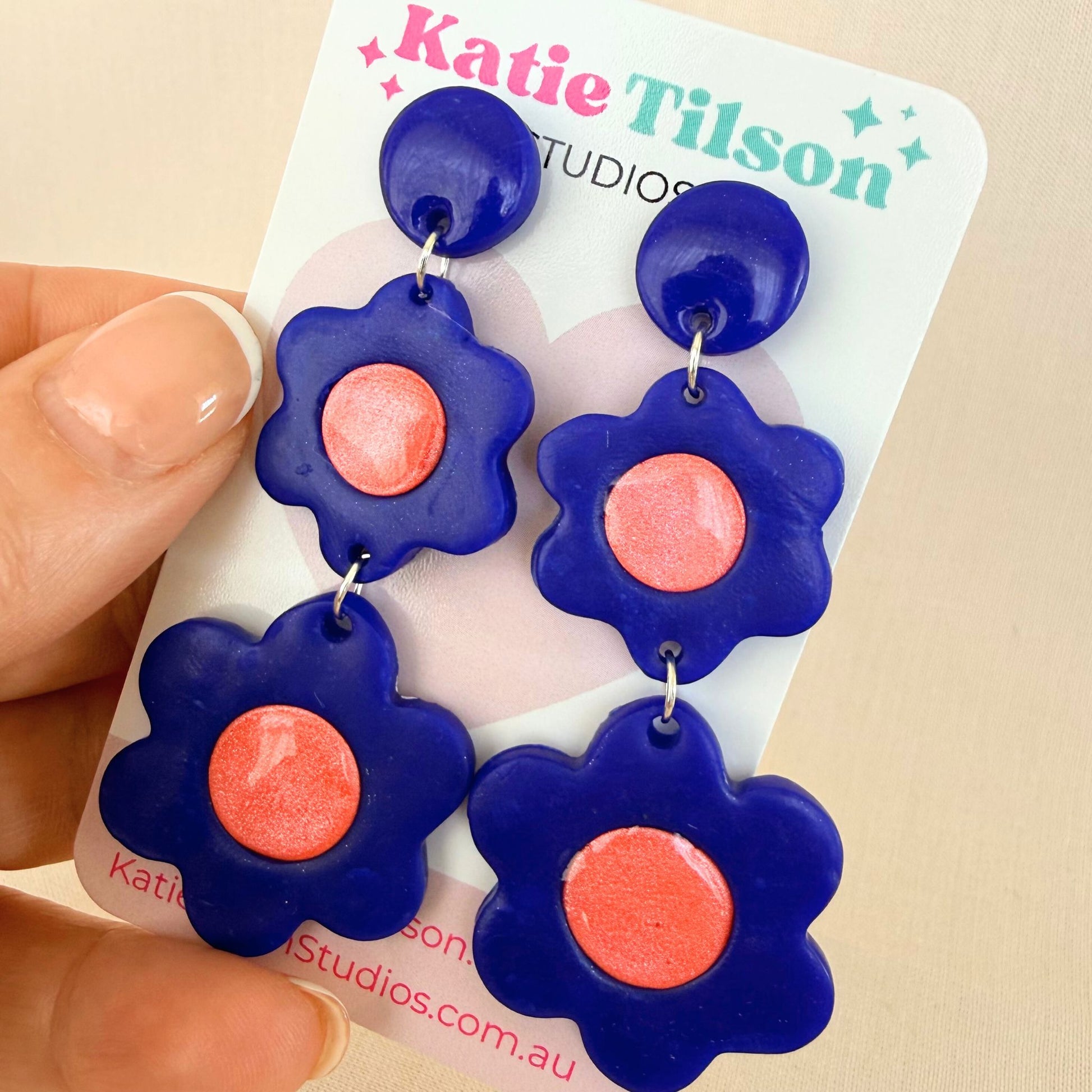 Flower-shaped dangle earrings with cobalt blue petals and coral centers on a Katie Tilson Studios card held by a hand.