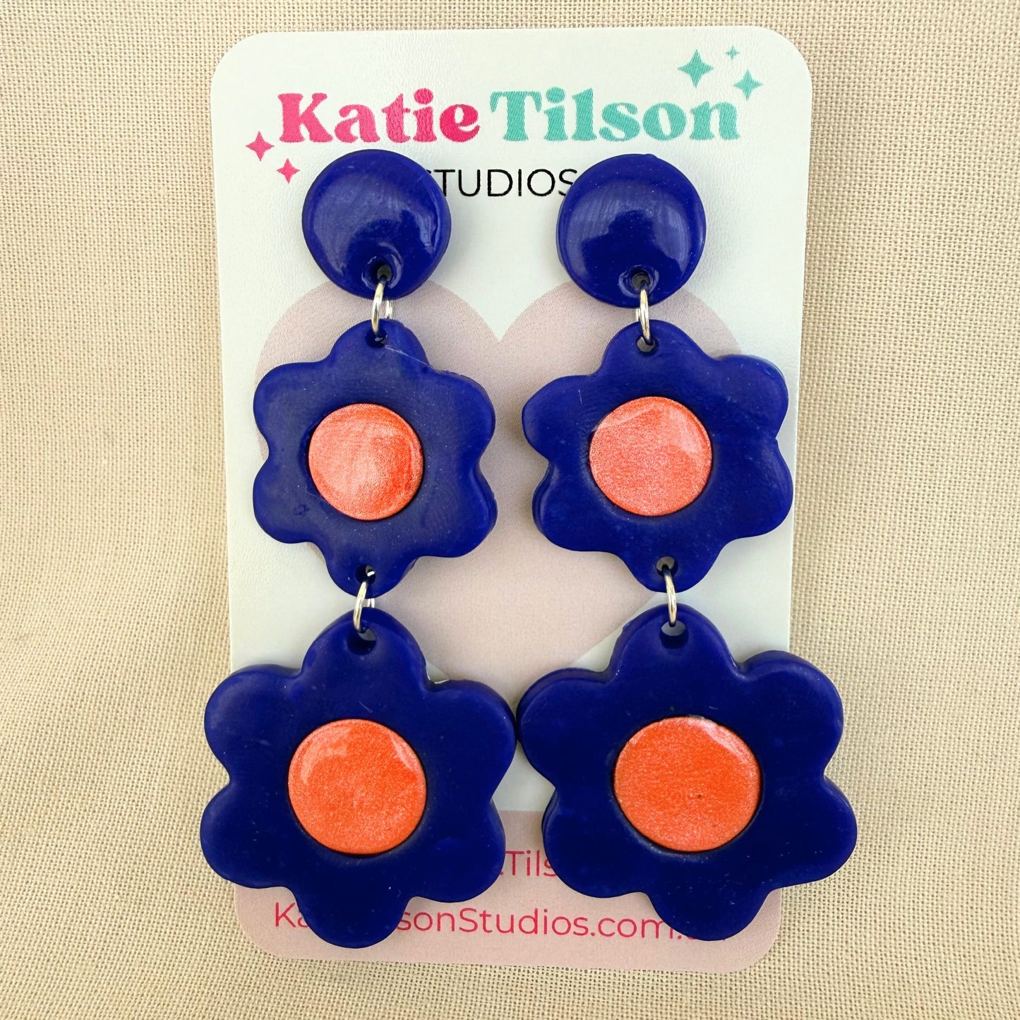 Flower-shaped dangle earrings with cobalt blue petals and coral centers on a Katie Tilson Studios card against a beige background.