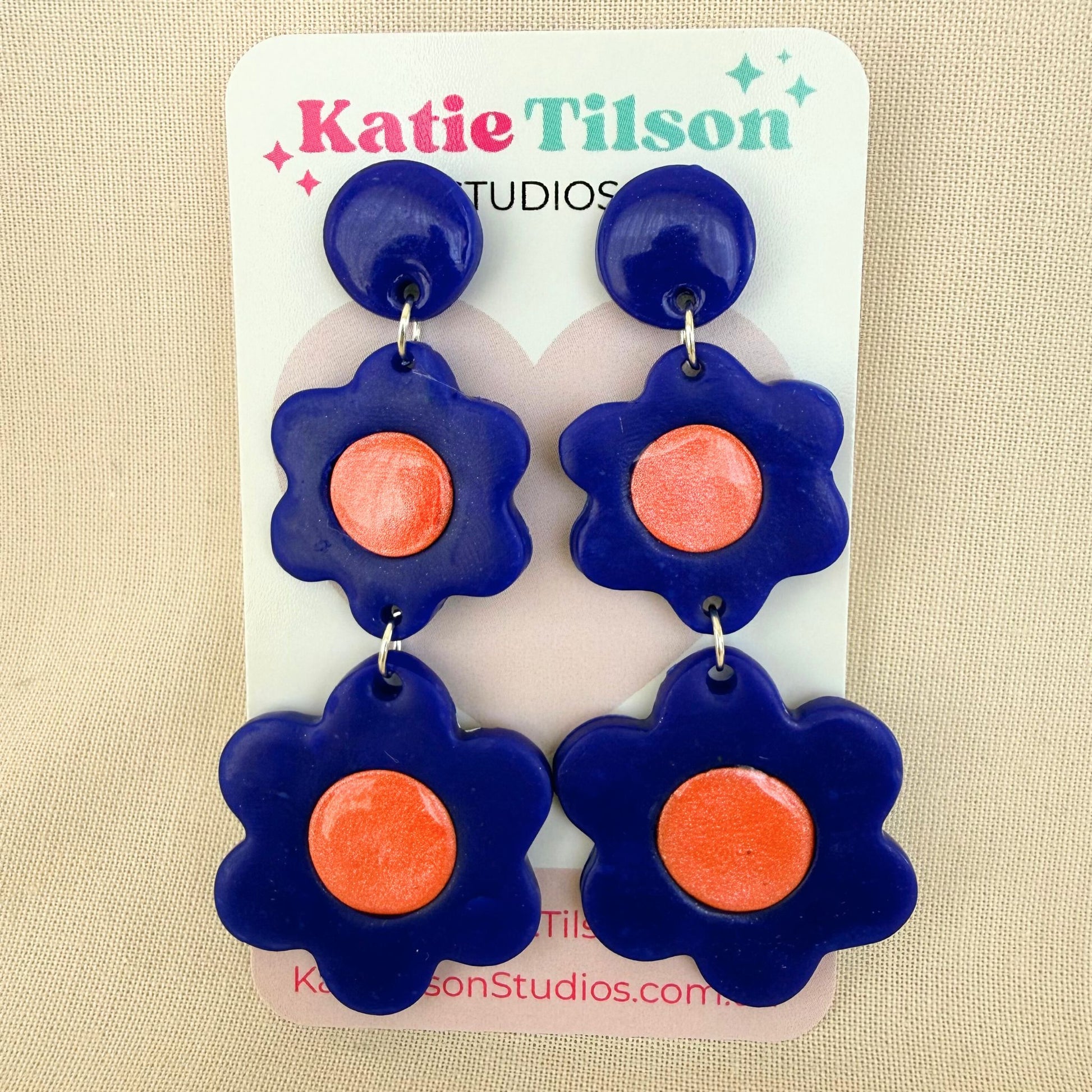 Flower-shaped dangle earrings with cobalt blue petals and coral centers on a Katie Tilson Studios card against a beige background.