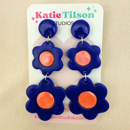 Flower-shaped dangle earrings with cobalt blue petals and coral centers on a Katie Tilson Studios card against a beige background.