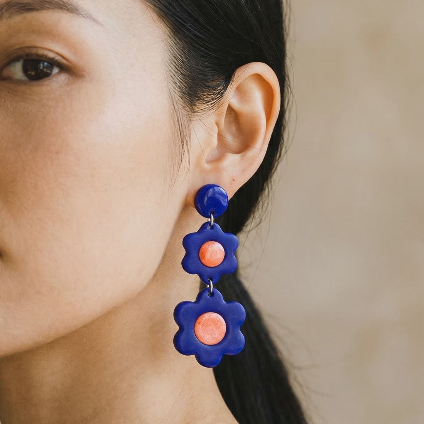 Close-up of a woman wearing cobalt blue 3 tier flower-shaped dangle earrings with glossy coral centers.