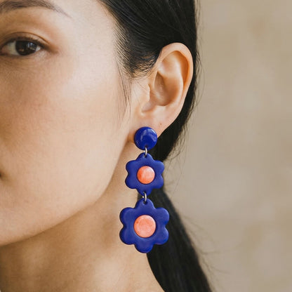 Close-up of a woman wearing cobalt blue 3 tier flower-shaped dangle earrings with glossy coral centers.