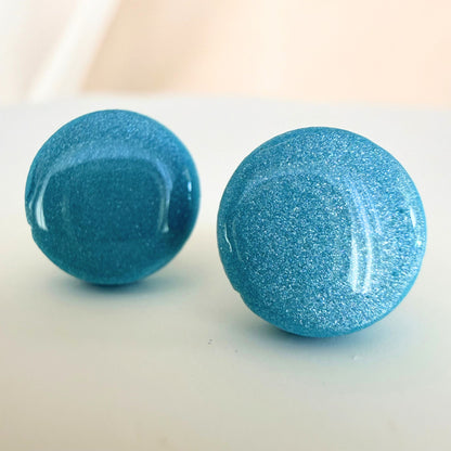 A pair of cornflower blue circle studs with shimmer and glossy resin finish resting on a white surface.