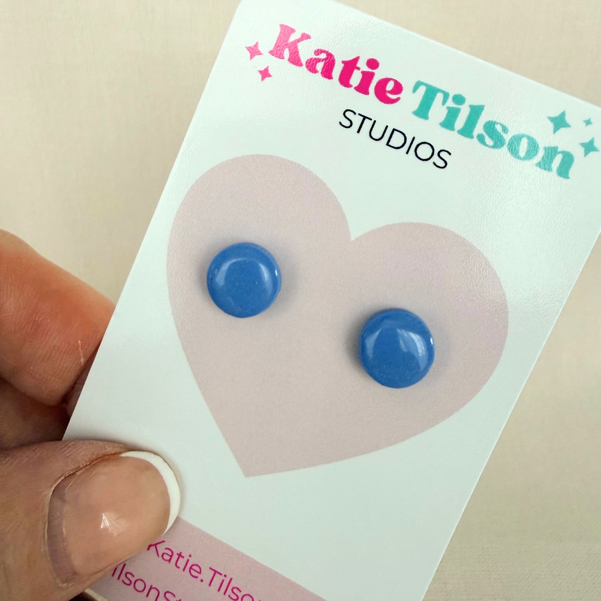 Cornflower blue circular stud earrings on a Katie Tilson Studios card held by a hand.