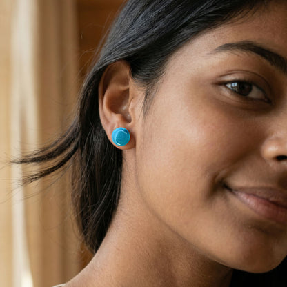 Close-up of a woman wearing round cornflower blue stud earrings with a glossy finish against a blurred background.