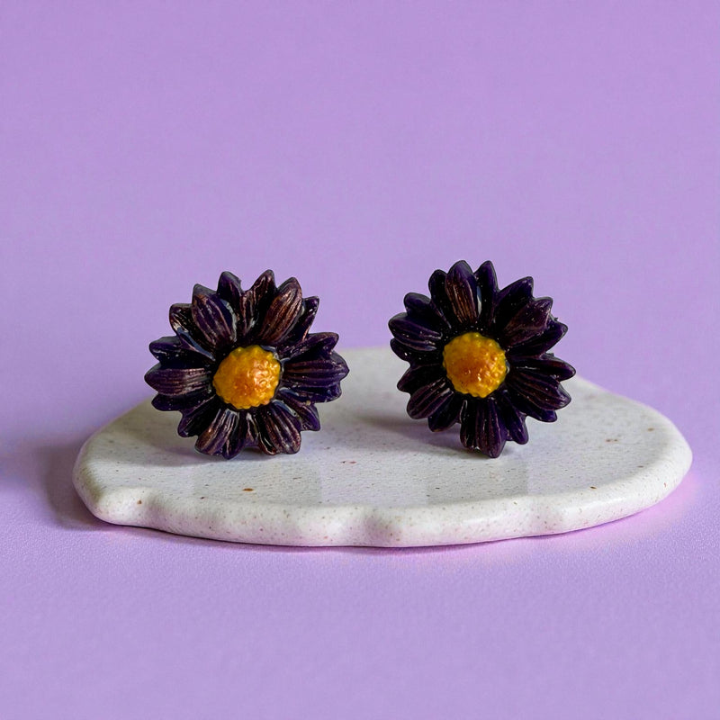 Purple daisy flower-shaped polymer clay stud earrings on a white stand against a purple background