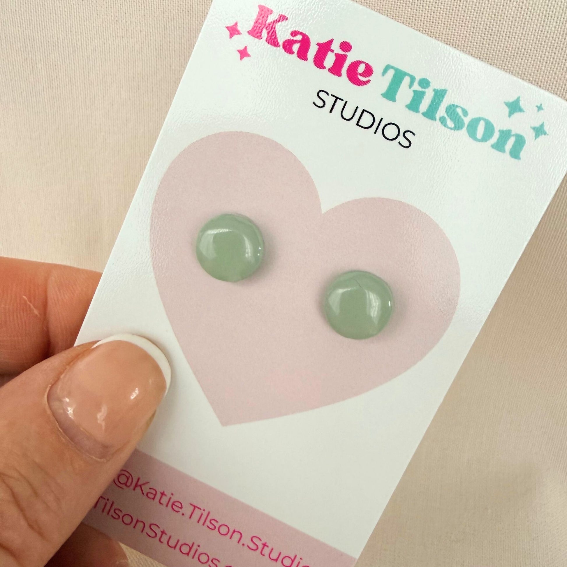 Eucalyptus green circular stud earrings on a Katie Tilson Studios card held by a hand.