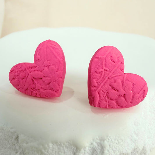 A pair of handmade stud earrings in pink heart design with embossed floral details resting on a white surface against a beige background.