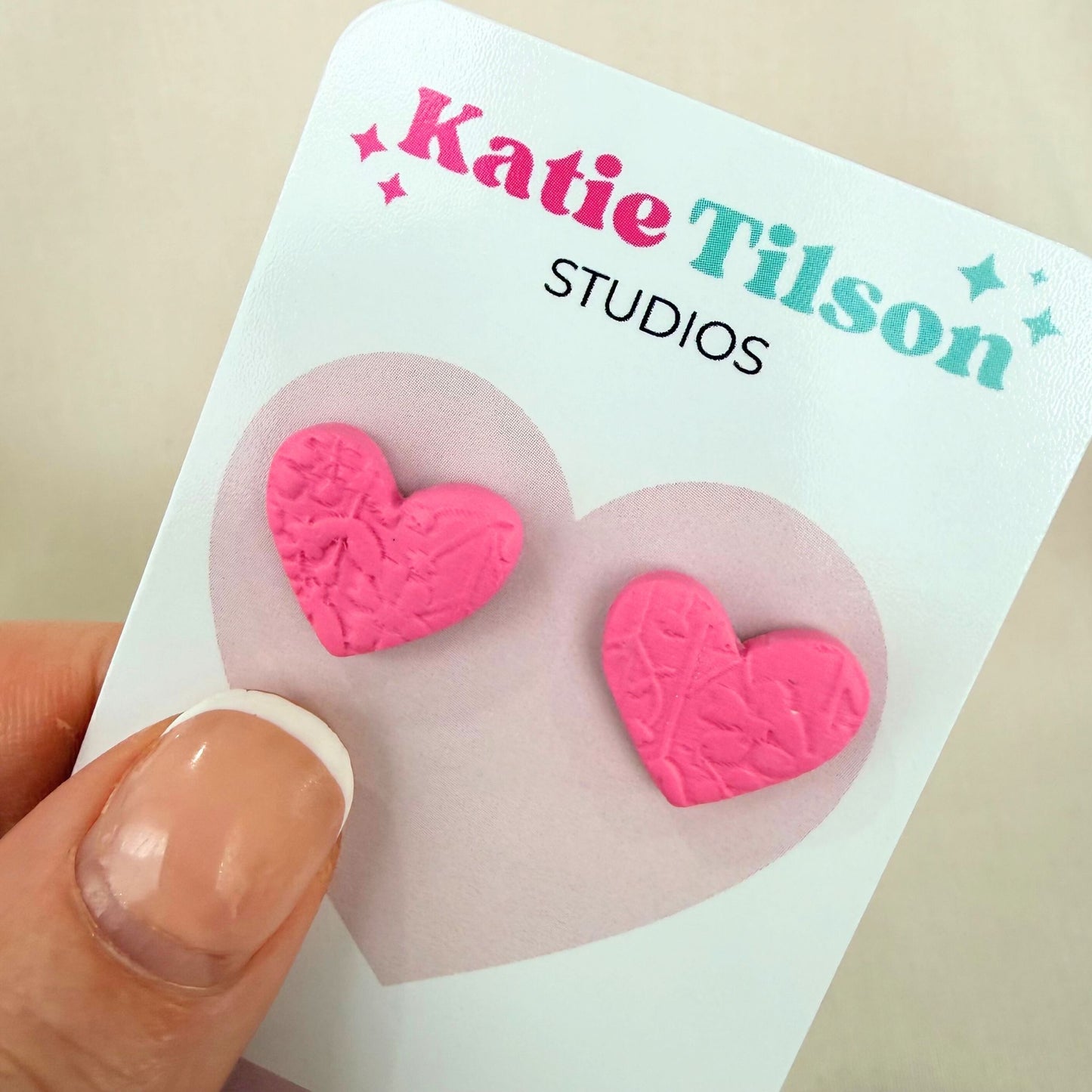 Pink heart-shaped stud earrings on a Katie Tilson Studios card held by a hand.