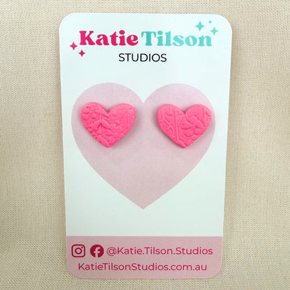 Pink heart shaped stud earrings with floral embossed texture on a Katie Tilson Studios branded earring card against a beige background.