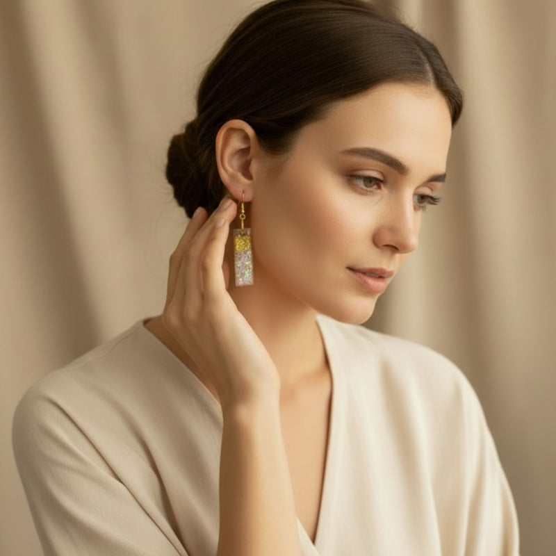 Woman wearing Australian handmade resin dangle earrings in a bar shape with gold leaf and sparkle against a beige background.