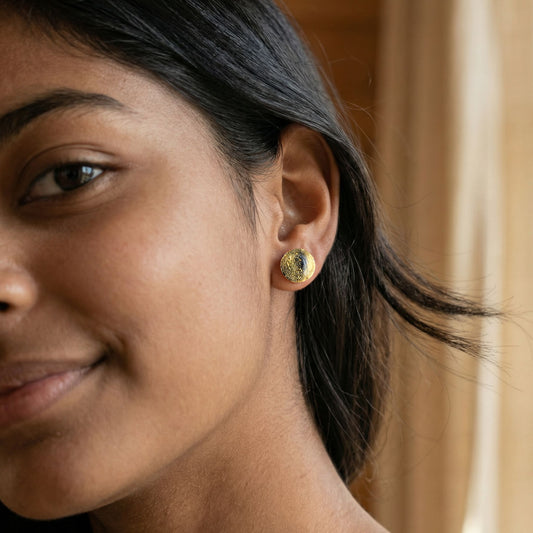 Close-up of a woman wearing round gold stud earrings with a glossy finish against a blurred background.