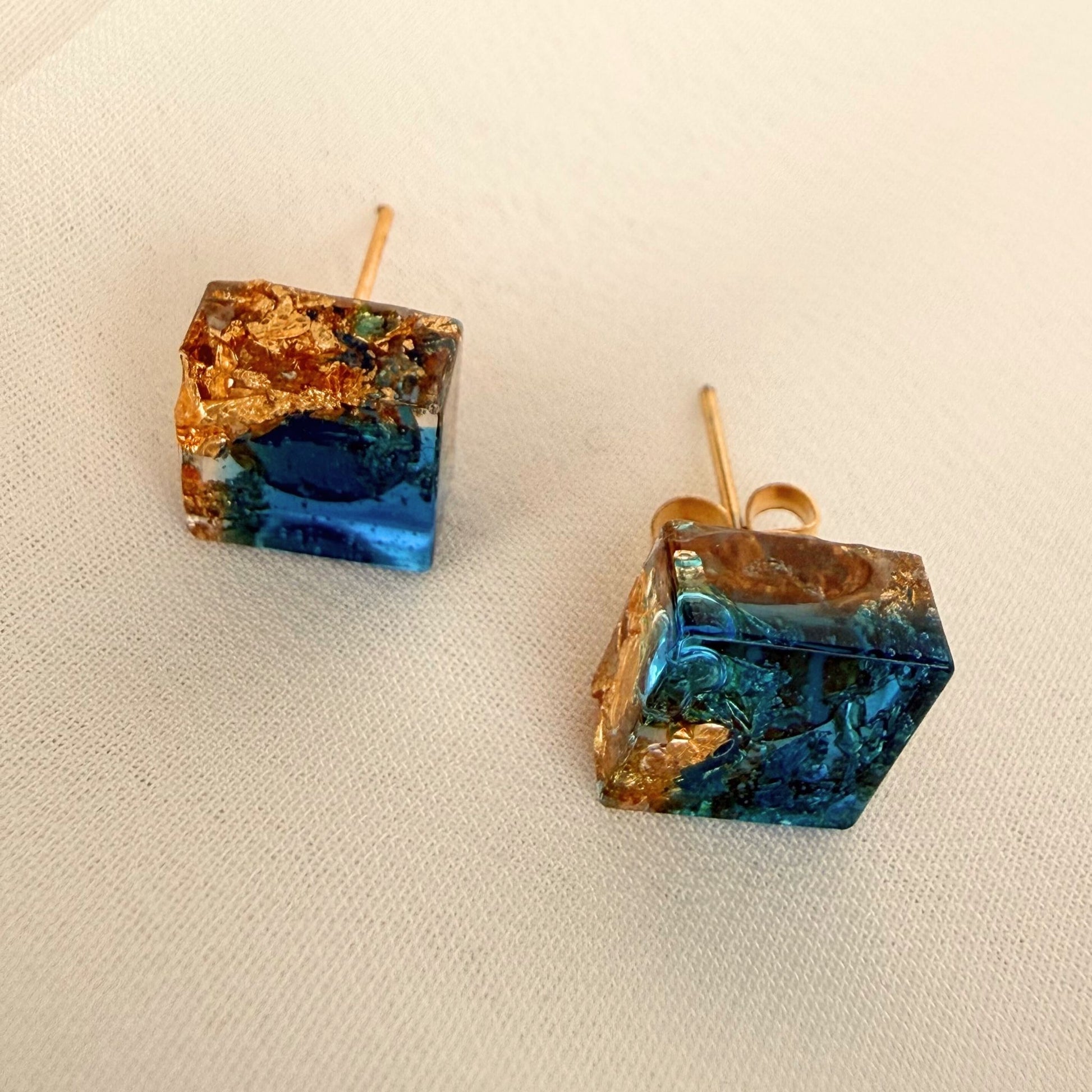 Gilded Tide handmade square resin stud earrings in teal and gold resting on white fabric.