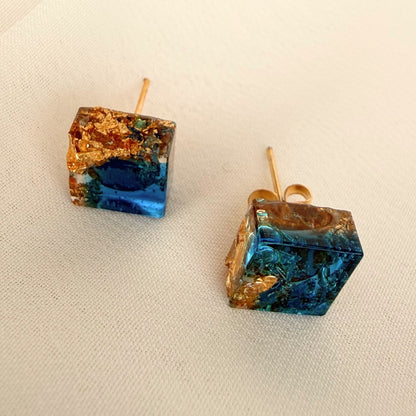 Gilded Tide handmade square resin stud earrings in teal and gold resting on white fabric.