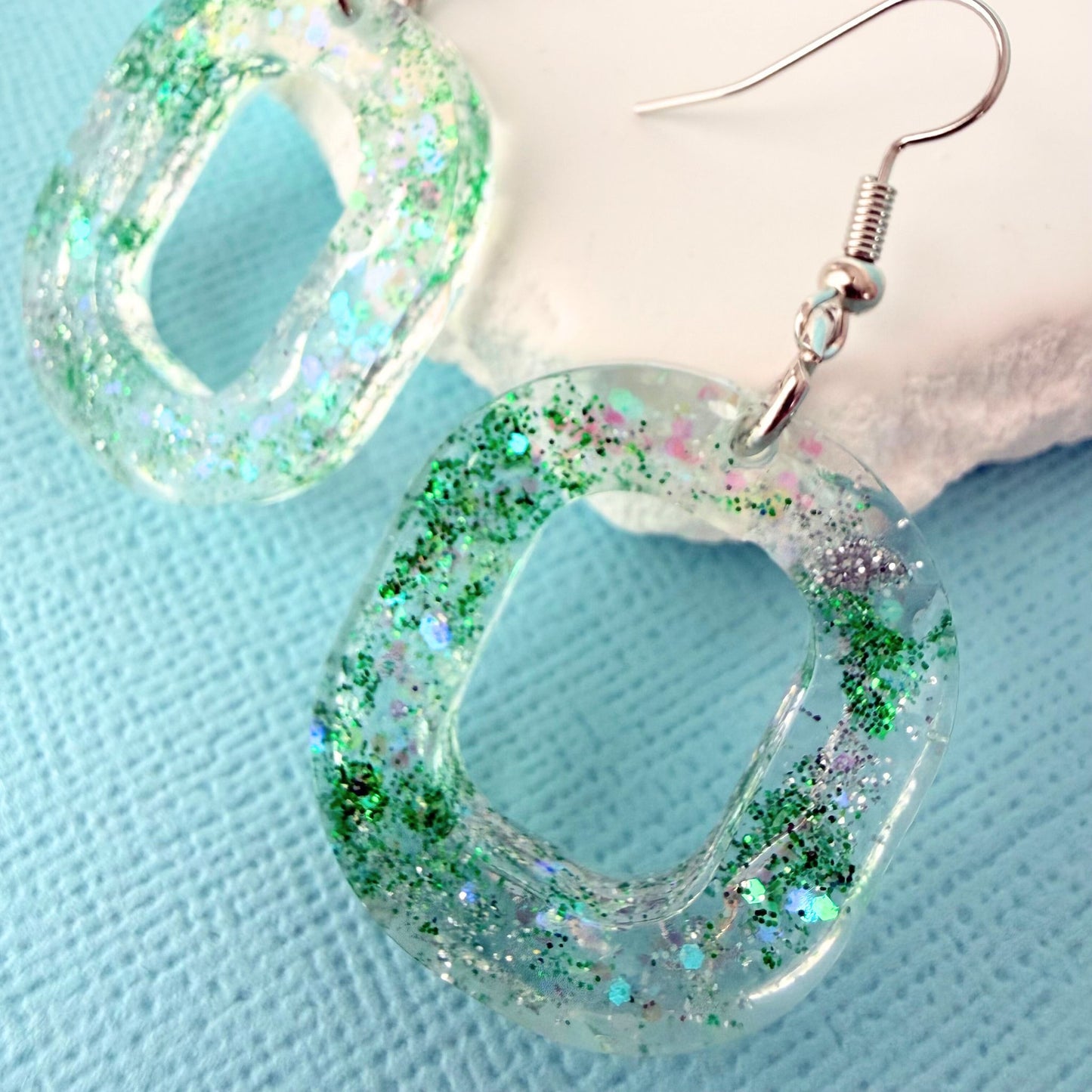 Close up of oval shaped resin glitter earrings with silver toned hooks resting on a white stone on a blue background.
