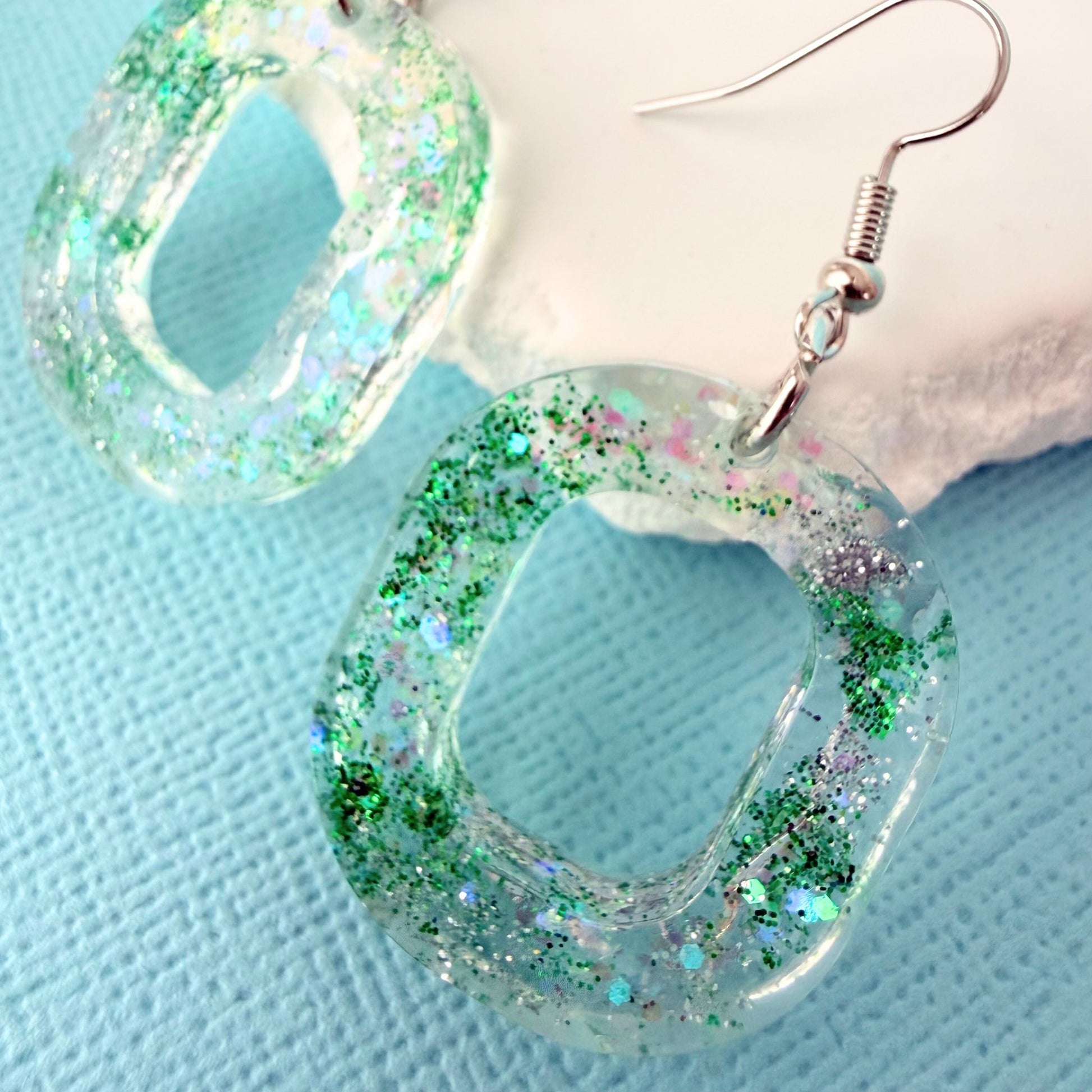 Close up of oval shaped resin glitter earrings with silver toned hooks resting on a white stone on a blue background.