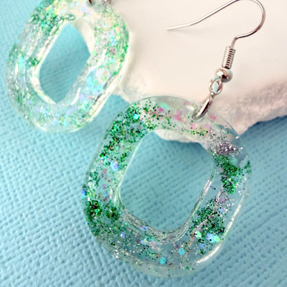 Close up of oval shaped resin glitter earrings with silver toned hooks resting on a white stone on a blue background.