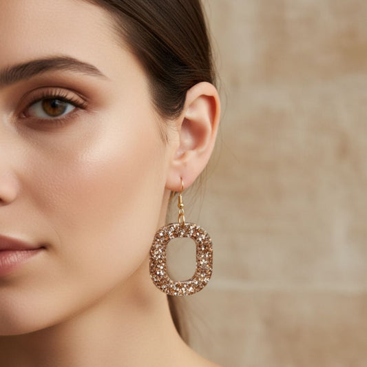 Close-up of a woman wearing sparkling gold and silver oval shaped dangle earring with gold toned hooks against a neutral background.