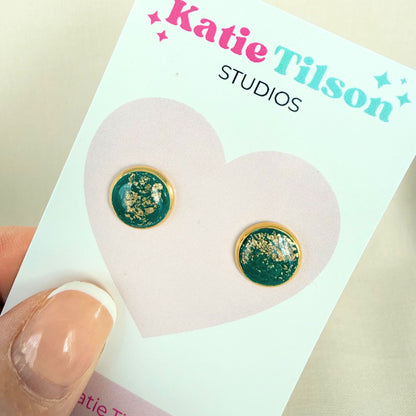 Emerald green and gold stud earrings on a Katie Tilson Studios card held by a hand.