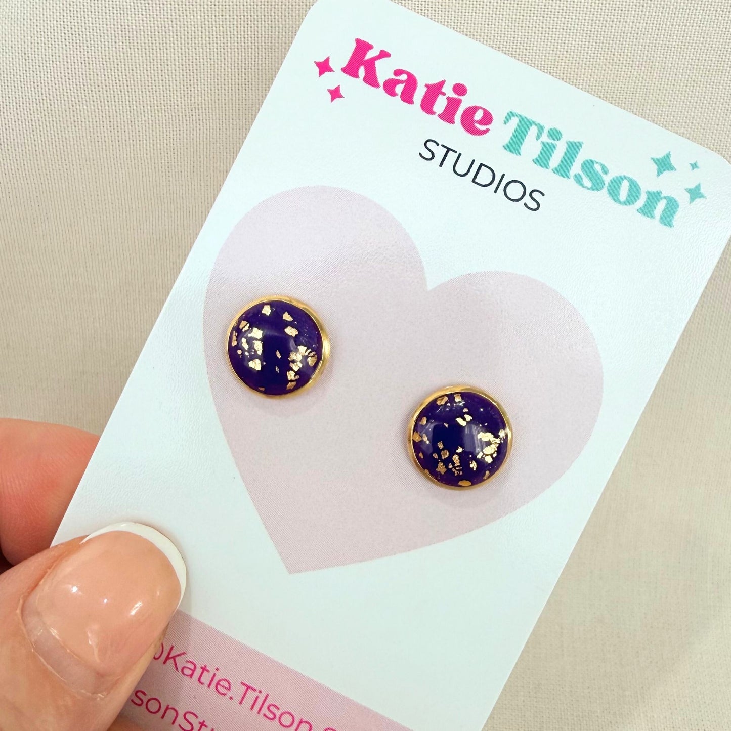 Purple stud earrings with gold details on a Katie Tilson Studios card held by a hand.
