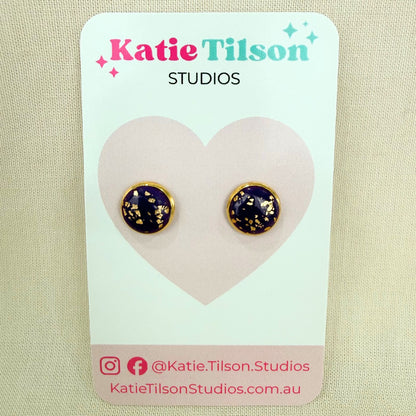 Purple handmade stud earrings with gold detail on a Katie Tilson Studios packaging with a beige background.