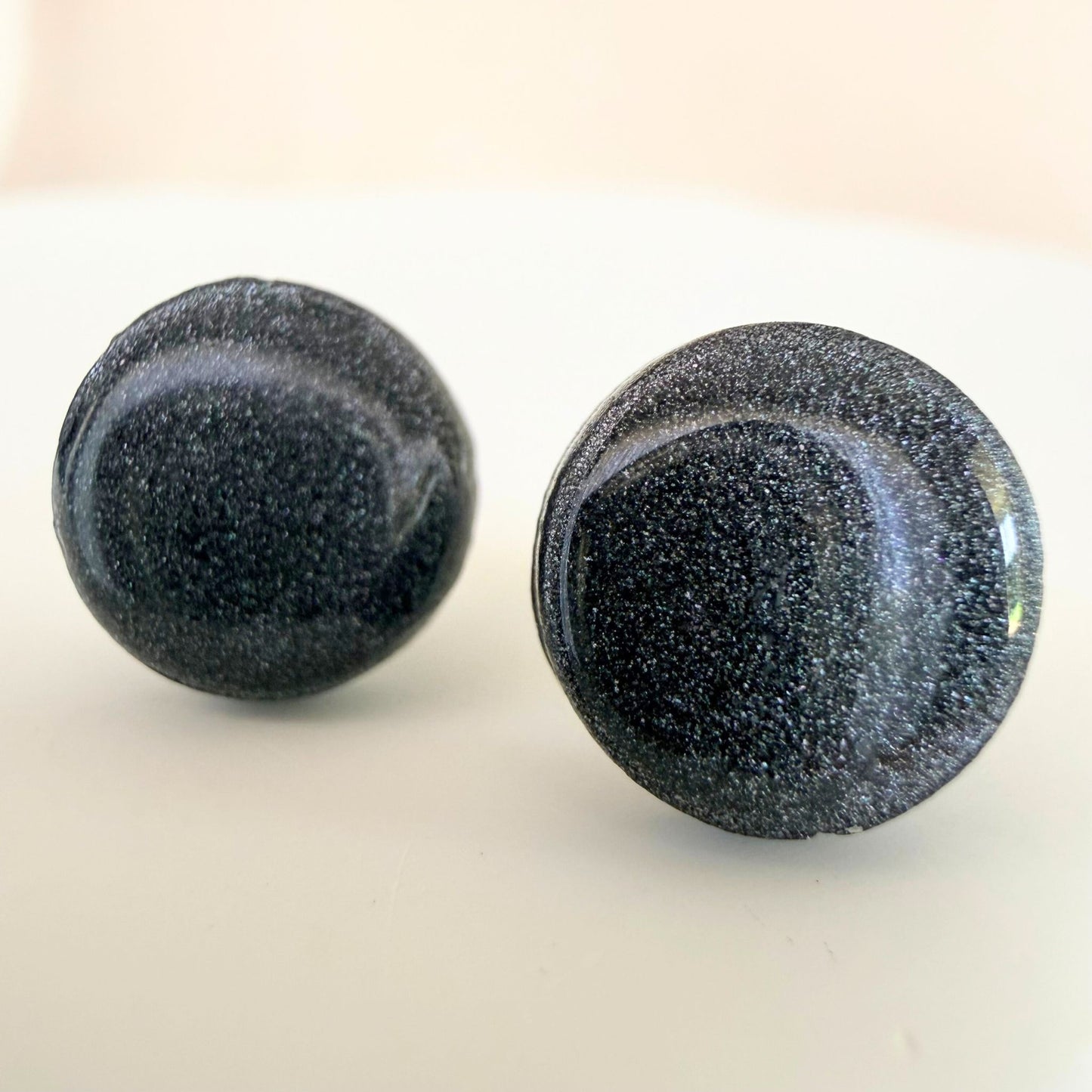A pair of graphite grey circle studs with shimmer and glossy resin finish resting on a white surface.