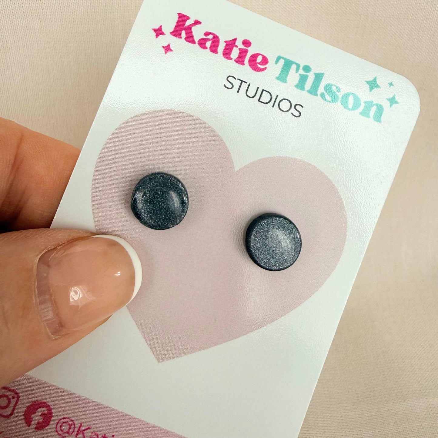 Graphite grey round stud earrings on a Katie Tilson Studios card held by a hand.