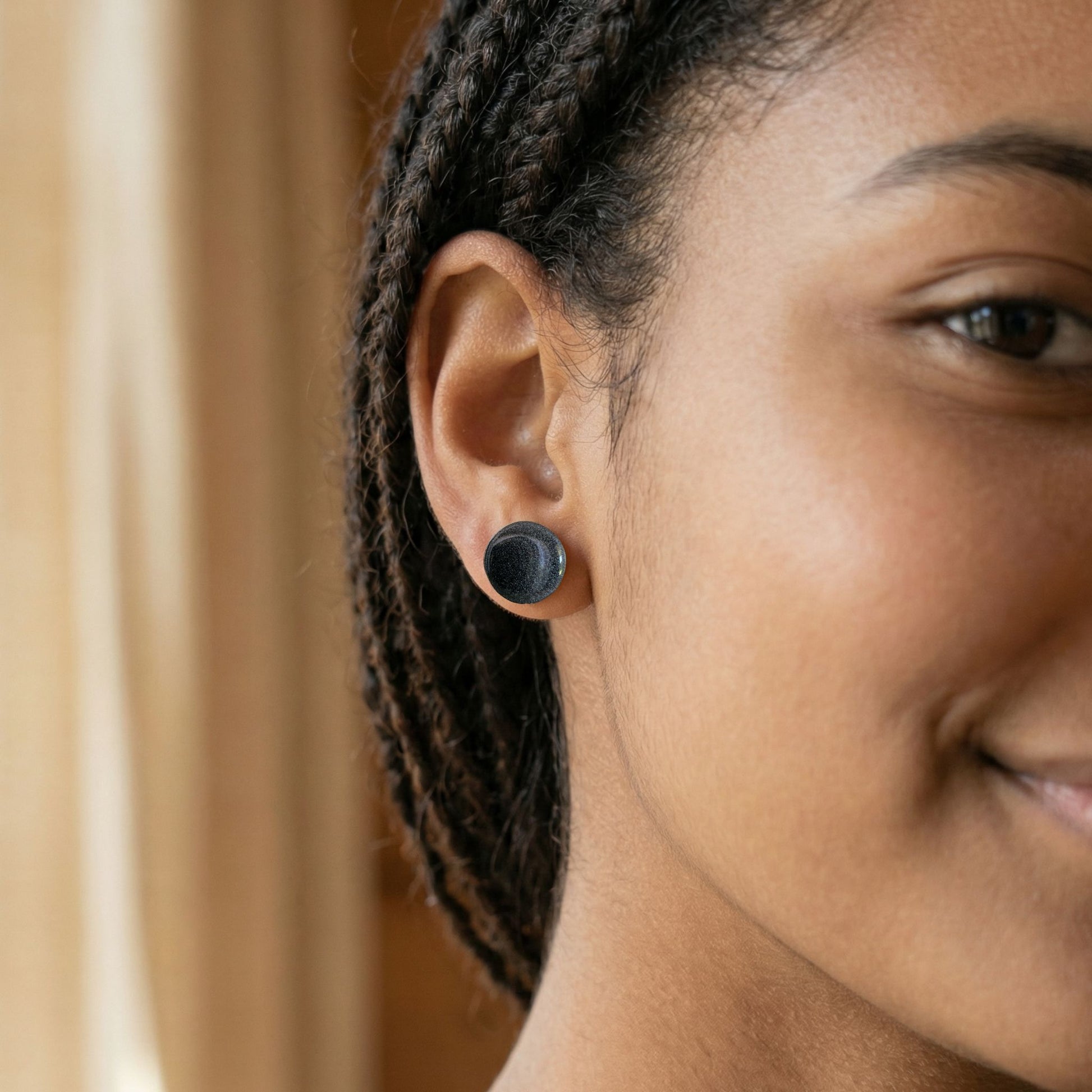 Close-up of a woman wearing round graphite grey stud earrings with a glossy finish against a blurred background.