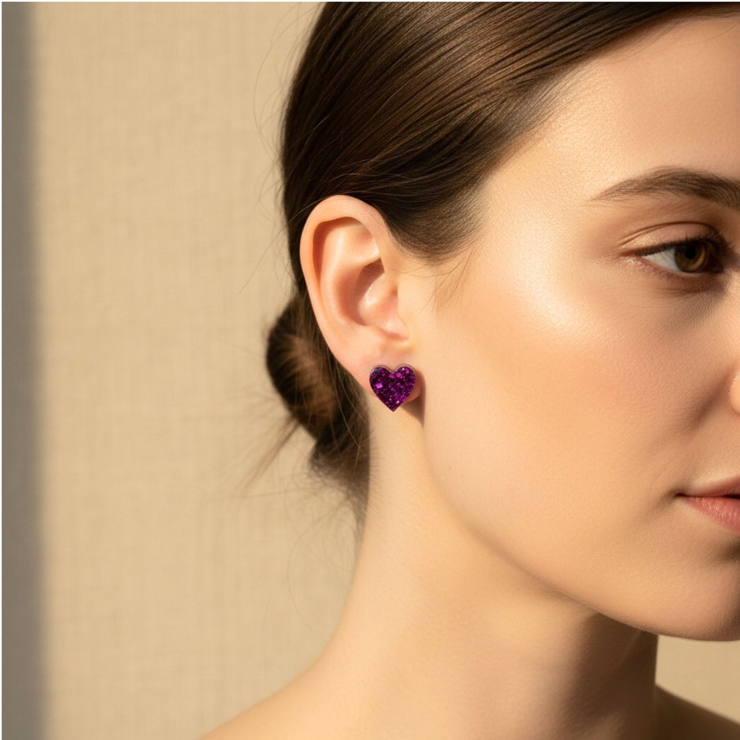 Close-up of a woman wearing purple sparkle heart-shaped resin earrings against a beige background.