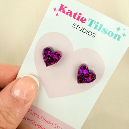 Purple resin heart-shaped stud earrings on a Katie Tilson Studios card held by a hand.