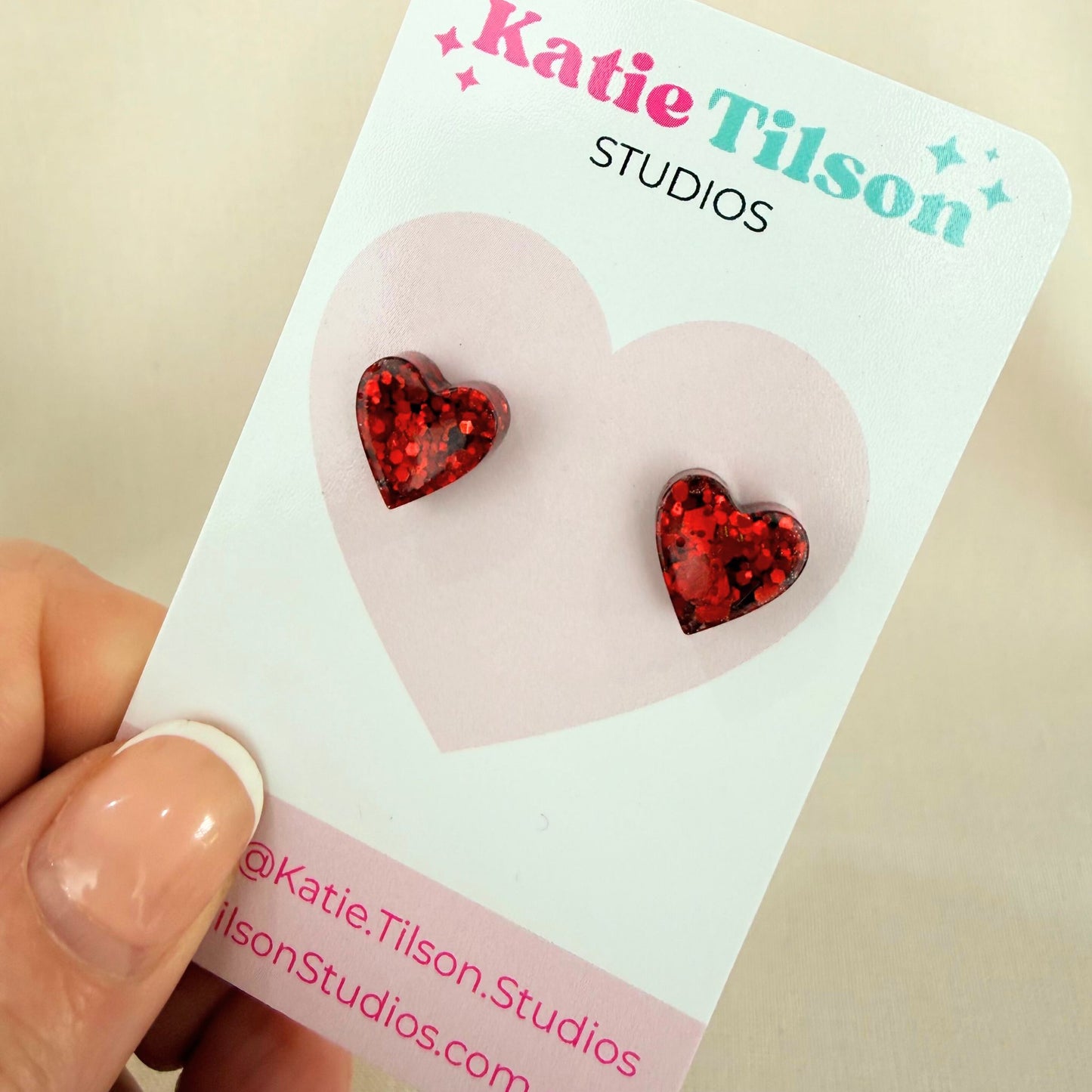 Red glitter resin heart-shaped stud earrings on a Katie Tilson Studios card held by a hand.
