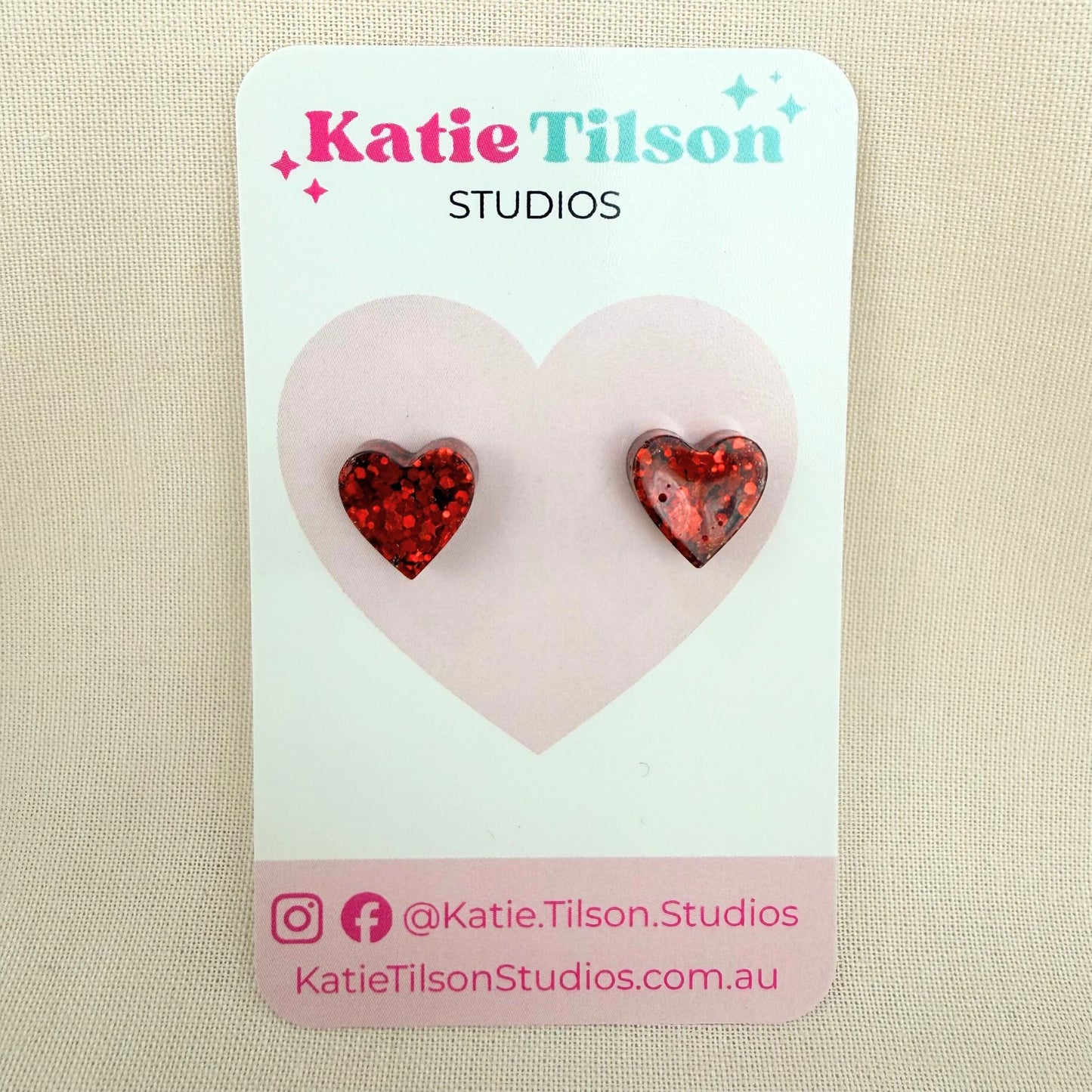 Red glitter resin heart-shaped stud earrings on a Katie Tilson Studios card against a beige background.
