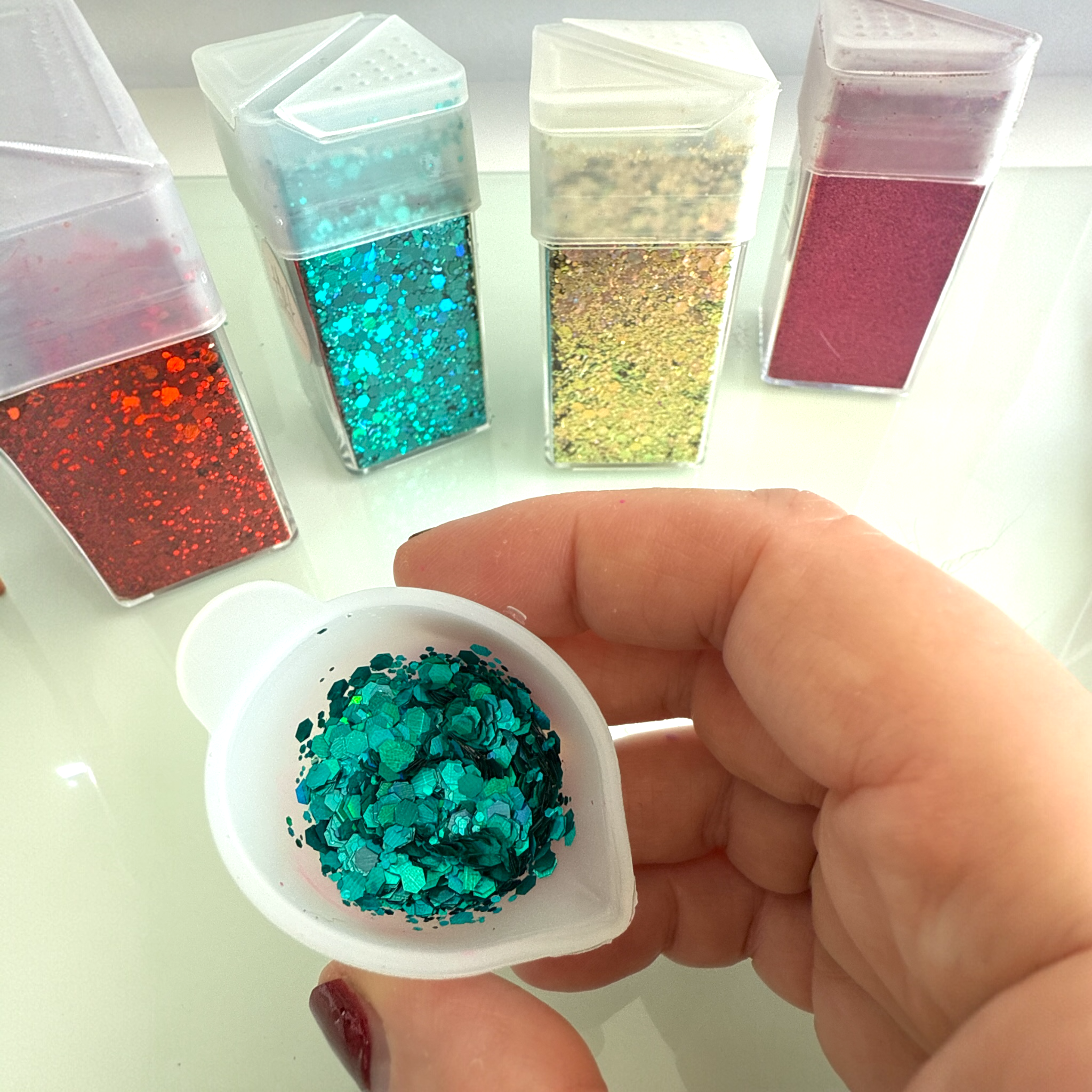 Hand holding a container of blue glitter with other glitter containers in the background.