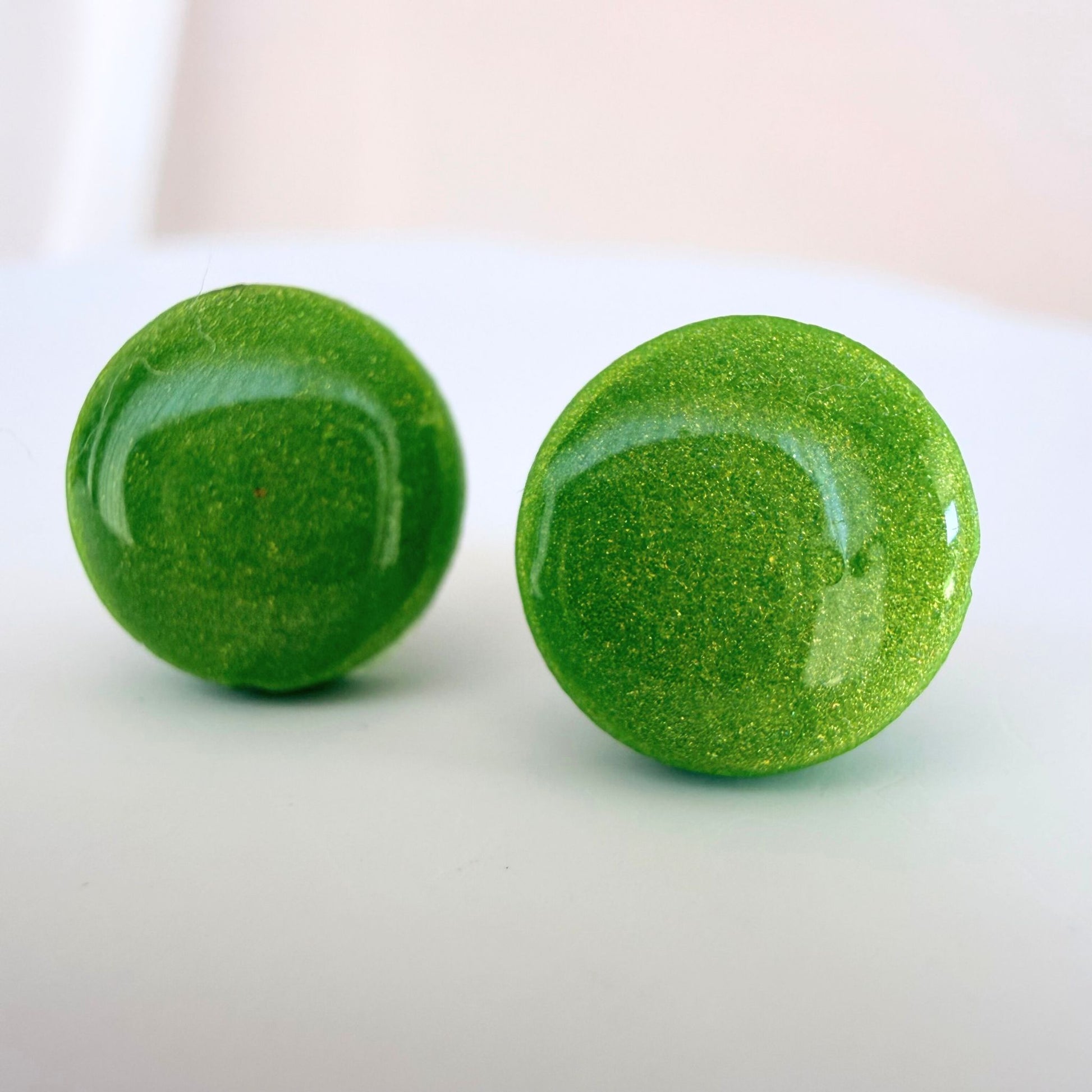 A pair of bright lime green circle studs with shimmer and glossy resin finish resting on a white surface.