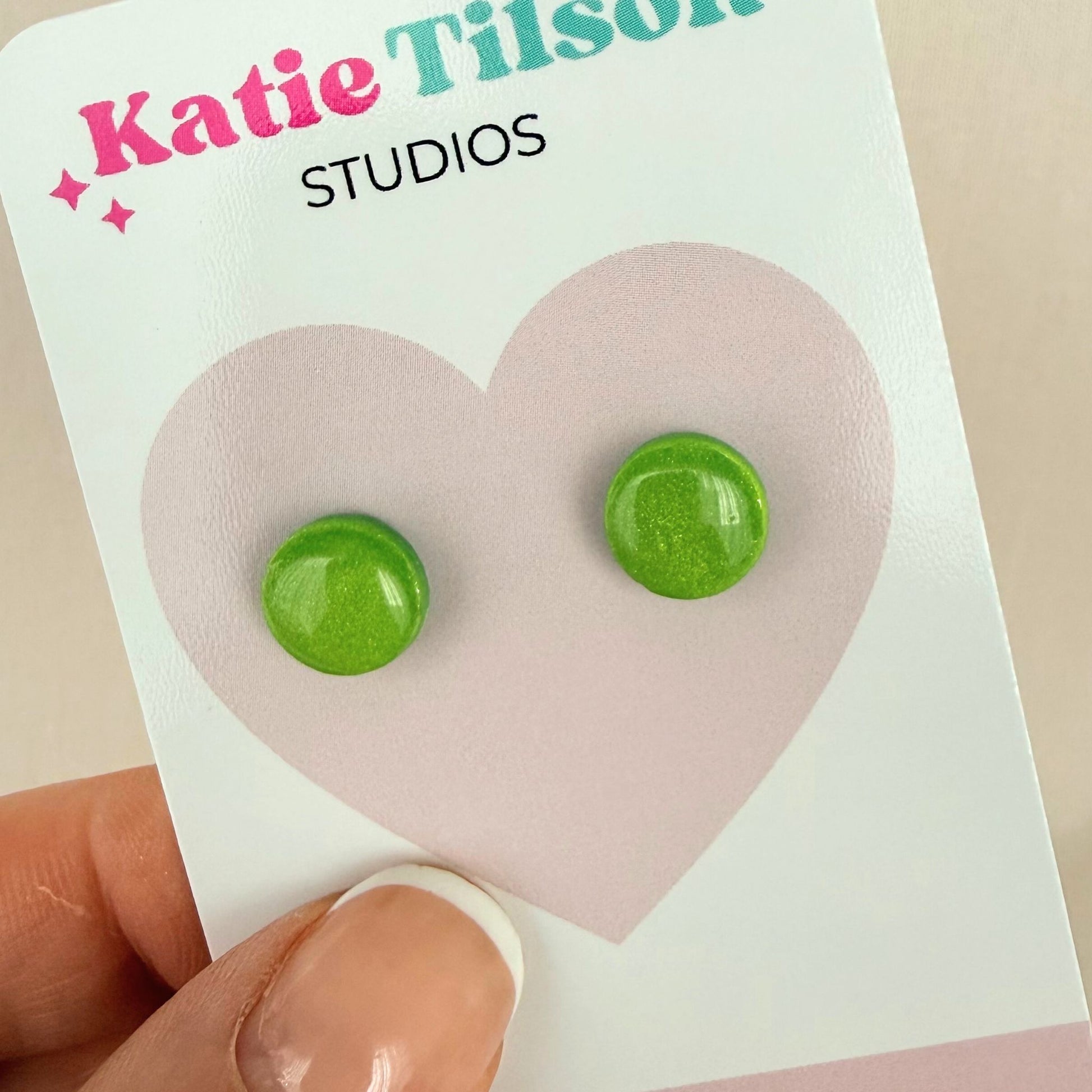 Lime green round stud earrings on a Katie Tilson Studios card held by a hand.